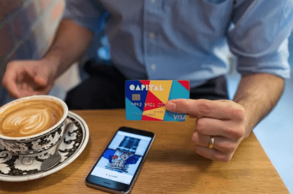 An image of a person holding a Qapital Visa Card while looking at their phone and drinking a cup of coffee.